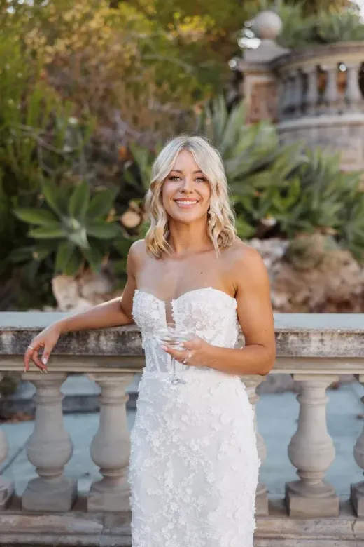 Bride in a beaded lace gown holding a glass at a garden balustrade — natural, luminous bridal makeup